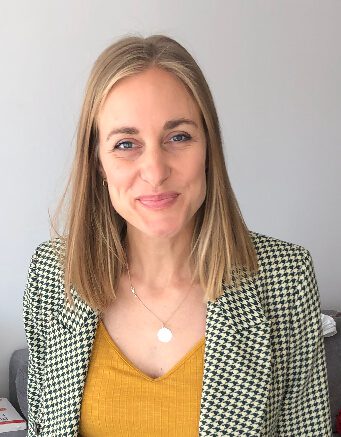 A white woman with shoulder-length blonde hair is smiling at the camera. She's wearing a yellow shirt, a green checkered blazer, and a gold necklace.