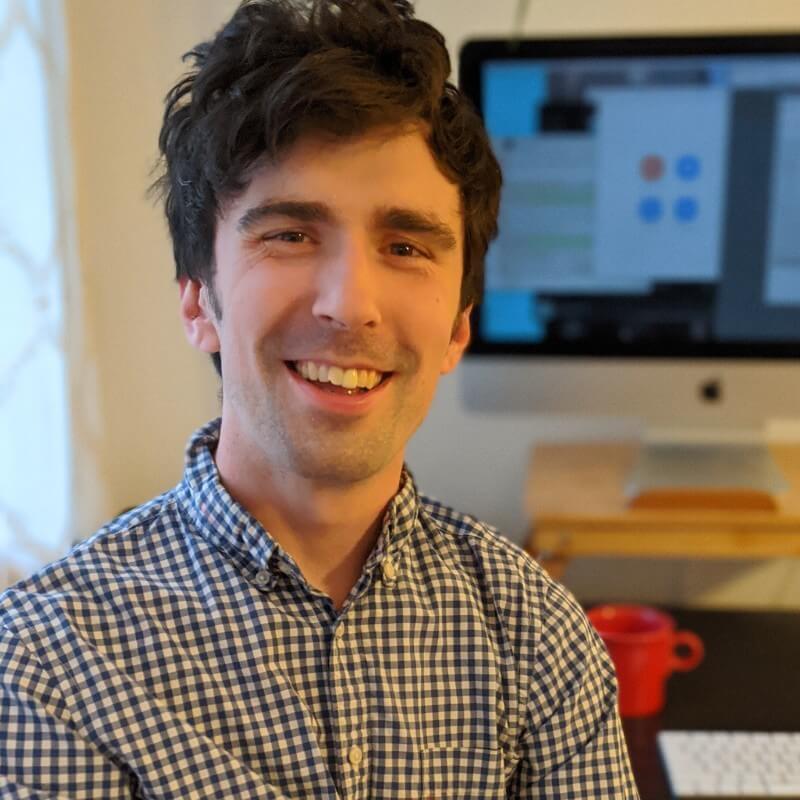 A white male with longer brown hair smiles. He is sitting in front of a computer and wearing a checkered shirt.