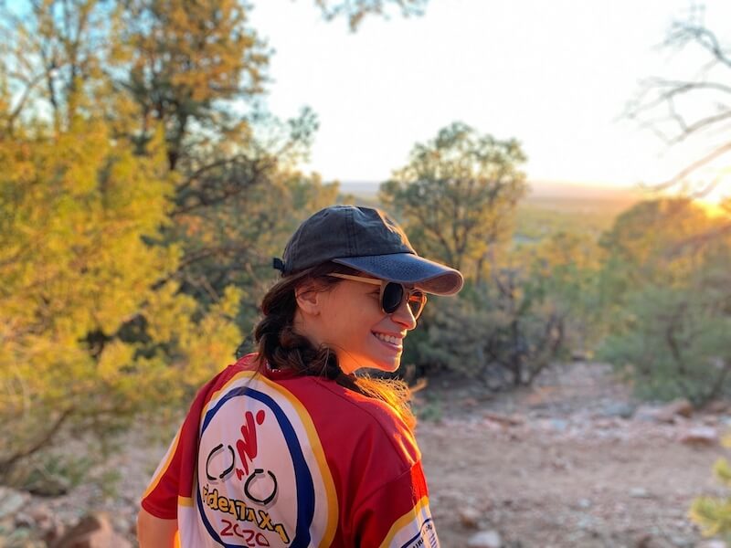A woman sits outside with her back to the camera but is turning her face toward the side. She is white with brown hair and wearing a blue baseball hat and a red shirt with a bike on the back.