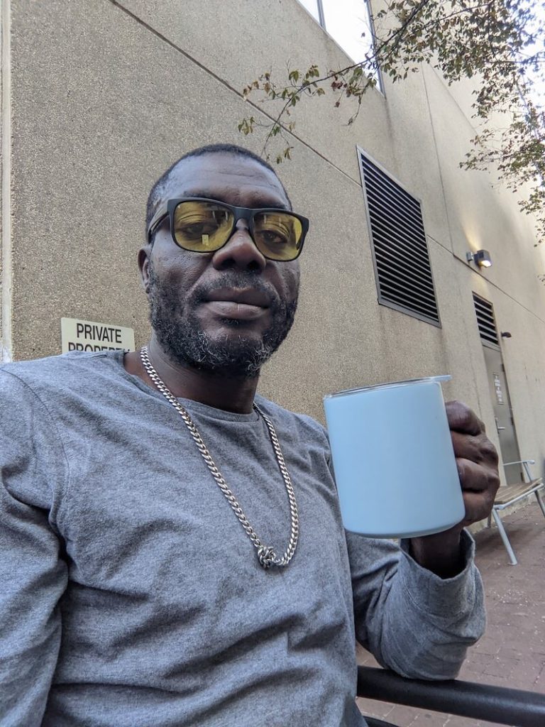 A black man with short hair and a trimmed beard, smiles at the camera. He is wearing a gray shirt and gold chain necklace and holding a travel mug.