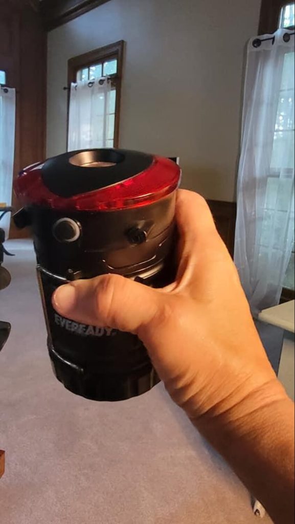 A person holding the closed camping lantern. The top of it is more visible, including the red parts for emergency flashing and the top for the flashlight.