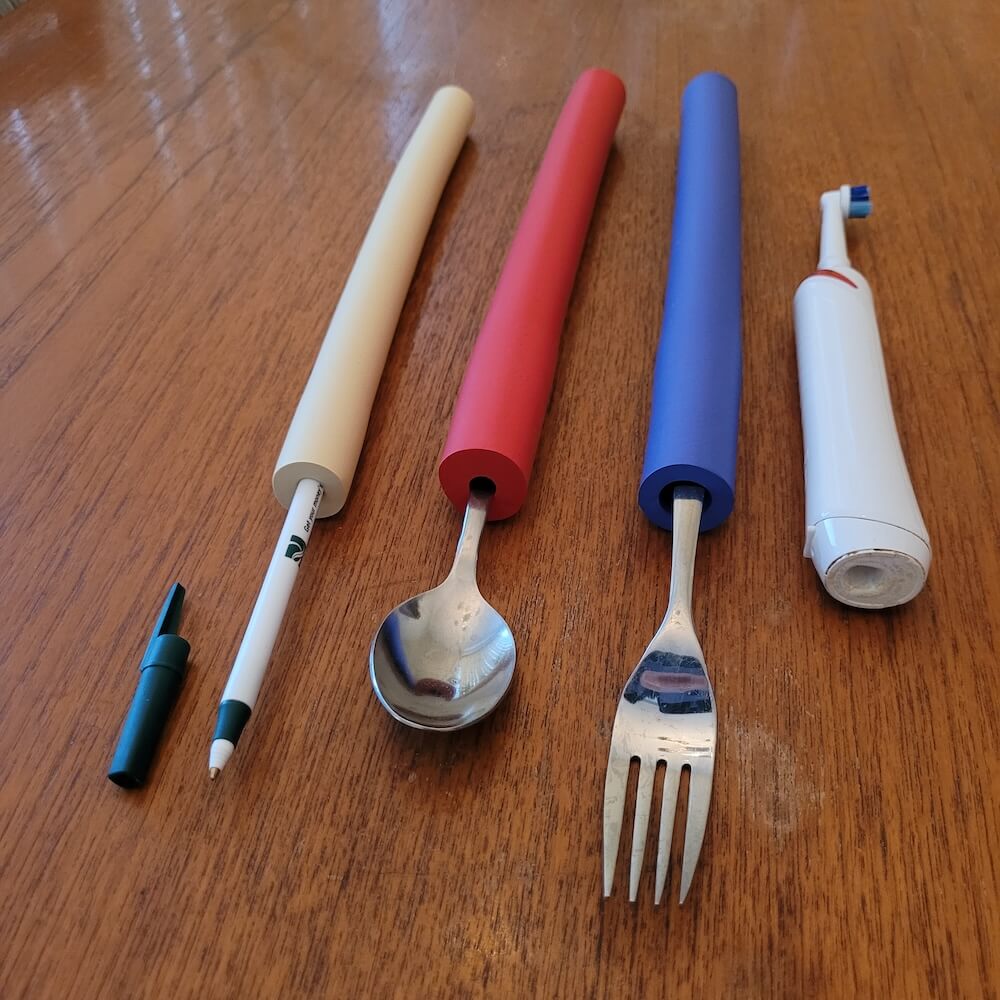 Vive foam tubes, a white one on a pen, a red one on a spoon, and a blue one on a fork. The grips do not fit on the electric toothbrush sitting on the table beside them.