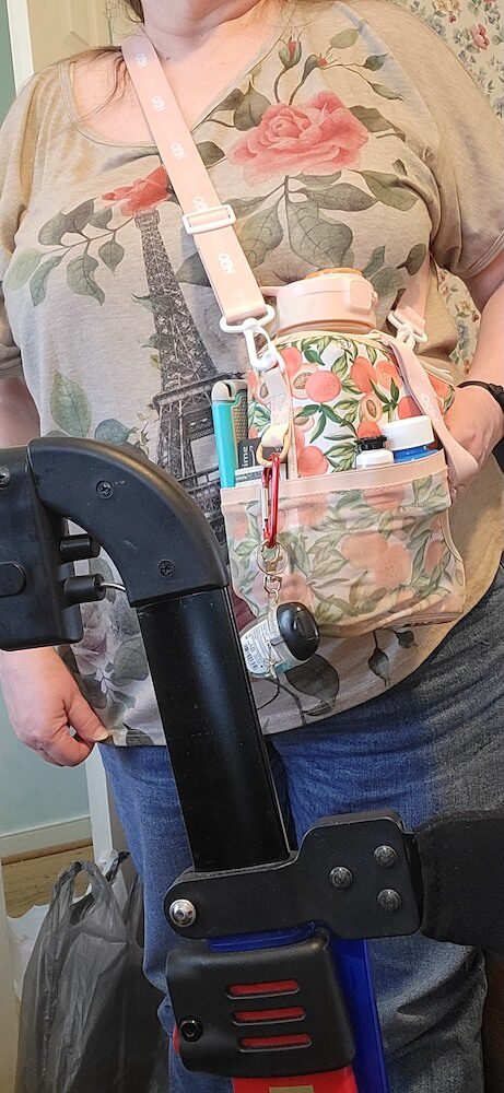 A person is wearing the Cube Water Bottle across their body. The pocket of the peach patterned water bottle is holding a phone, money, lotion and more. Hand sanitizer is clipped to the bottle.