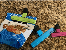 A green clip with black rubber on the handle is holding a chip bag closed. Blue and pink clips in different sizes are sitting beside the bag.