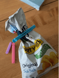 A blue clip has been snapped across the top of chip bag that has been pulled together at the top. Pink and purple clips of different sizes are on the table beside the bag.